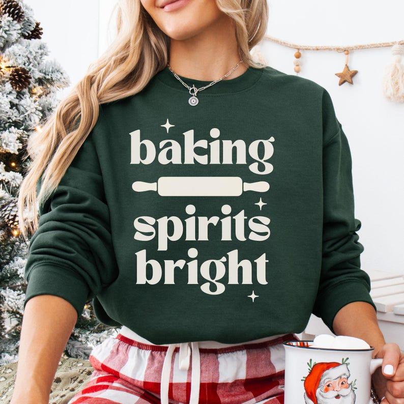 K&ouml;nnte beinhalten: Ein dunkelgr&uuml;nes Langarm-Sweatshirt mit dem cremefarbenen Schriftzug "baking spirits bright", einer Nudelholz-Grafik und kleinen Sternakzenten, die mittig auf der Vorderseite platziert sind.