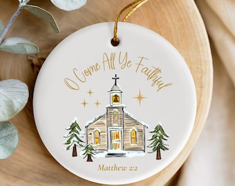 May include: A round, white Christmas ornament with the text "O Come All Ye Faithful" in gold script. The ornament features a church illustration with pine trees and the text "Matthew 2:2". A gold string is attached for hanging.