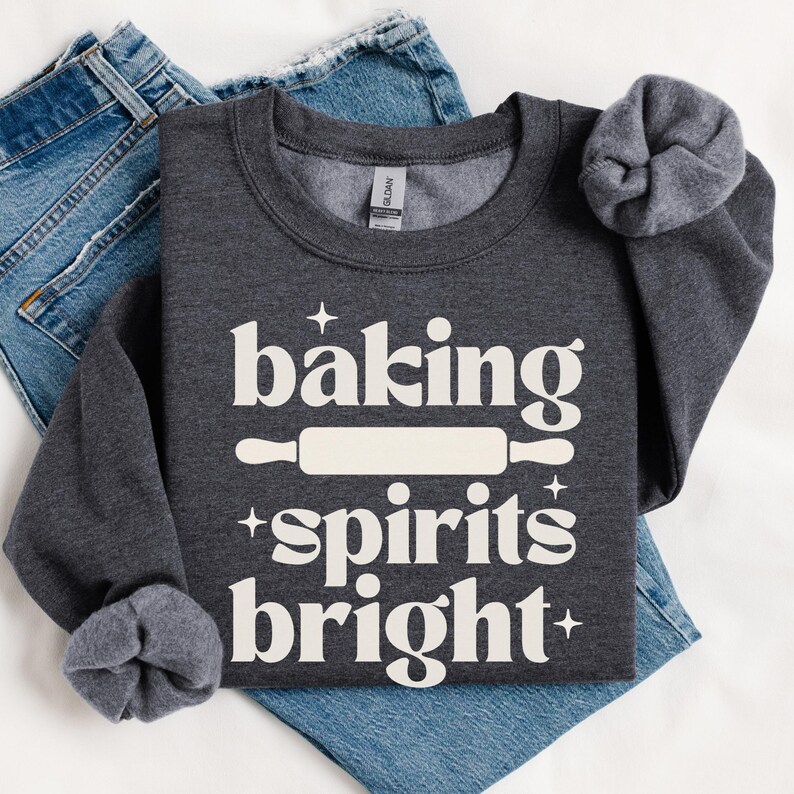 K&ouml;nnte beinhalten: Ein dunkelgraues Sweatshirt mit Rundhalsausschnitt und dem wei&szlig;en Schriftzug "baking spirits bright", einer Nudelholz-Grafik in der Mitte und kleinen Sternakzenten. Das Sweatshirt liegt auf einer blauen Jeans.