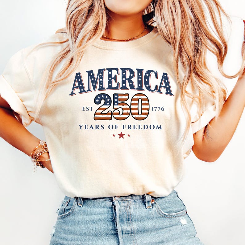 May include: Cream-colored t-shirt with the text "AMERICA 250 YEARS OF FREEDOM" in navy blue and red. The number 250 is designed with a red and white striped pattern, and the shirt has short sleeves.