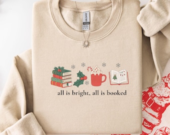 Könnte beinhalten: Beiges Sweatshirt mit Rundhalsausschnitt und dem Text "all is bright, all is booked". Die Grafik zeigt einen Stapel Bücher, Stechpalmen, eine Tasse heiße Schokolade und ein offenes Buch mit einem Weihnachtsbaum.
