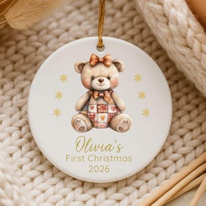 May include: A round white ceramic ornament featuring a brown teddy bear with a plaid heart-patterned belly, a bow on its head, and a bow tie. Gold snowflakes surround the bear. The text reads Olivia's First Christmas 2026 in gold lettering.