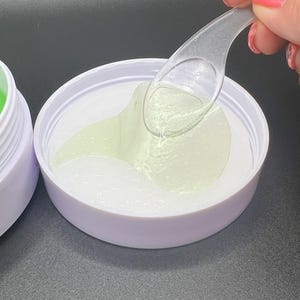 May include: A white jar with a light purple rim containing eye patches with a light green gel. A clear plastic spoon is removing a patch. The background is dark.