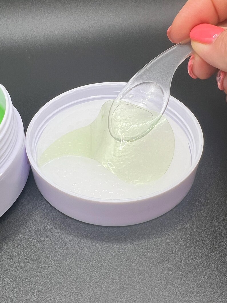 May include: A white jar with a light purple rim containing eye patches with a light green gel. A clear plastic spoon is removing a patch. The background is dark.