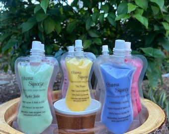 May include: A display of Aroma Squeeze wax melts in various colors, including green, yellow, blue, and pink. Each pouch is labeled with a scent name and details. The products are arranged on a wooden surface.
