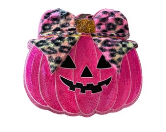 May include: A bright pink sequined pumpkin patch featuring a black felt jack-o'-lantern face with triangular eyes and a jagged mouth. A large bow with a pink, black, and tan leopard print pattern sits on top, accented by a small gold glitter center.