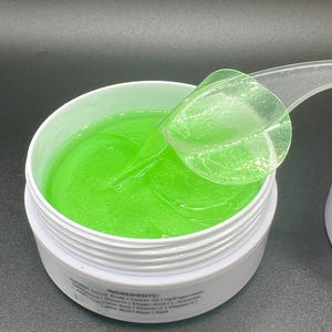 May include: A white jar filled with bright green gel, with a clear plastic scoop partially submerged. The gel has a smooth, reflective surface. The jar's label lists ingredients. The background is a dark grey.