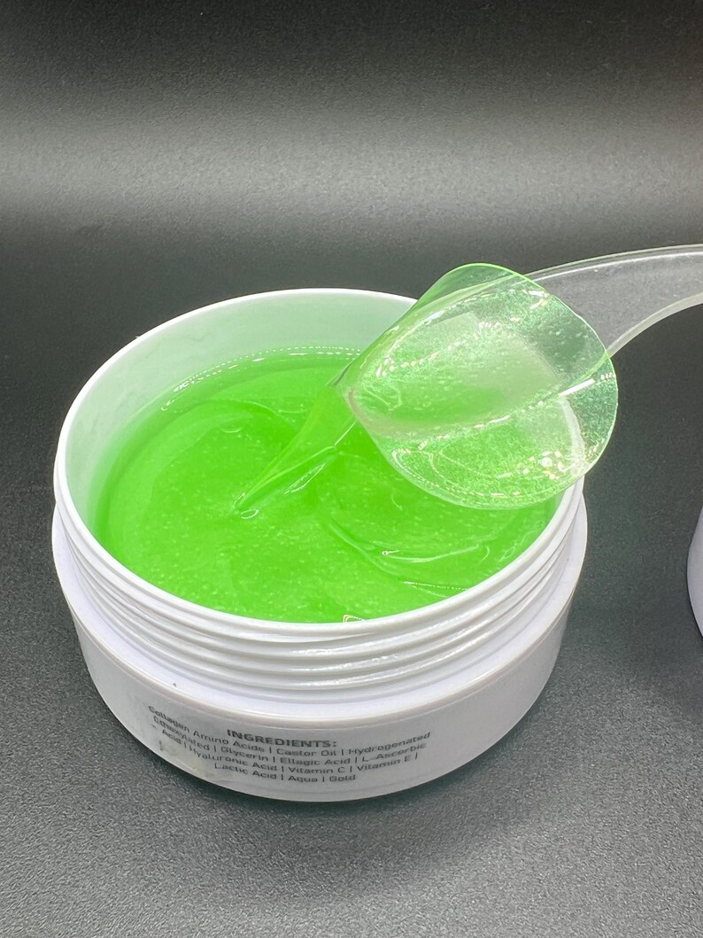 May include: A white jar filled with bright green gel, with a clear plastic scoop partially submerged. The gel has a smooth, reflective surface. The jar's label lists ingredients. The background is a dark grey.