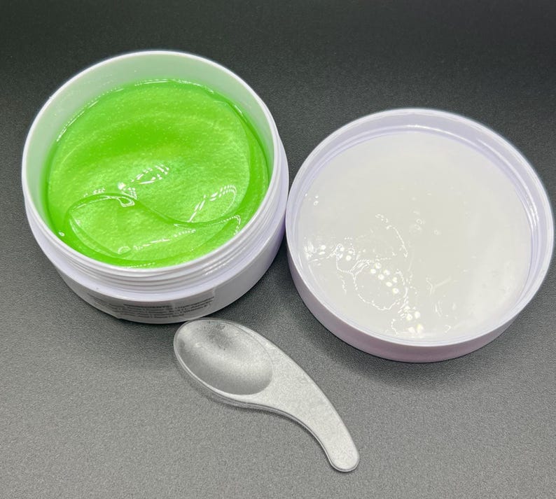 May include: Open white jar with bright green gel eye masks, a white lid, and a small silver spoon. The eye masks have a smooth, shiny texture. The lid is next to the jar. The spoon is on the bottom.