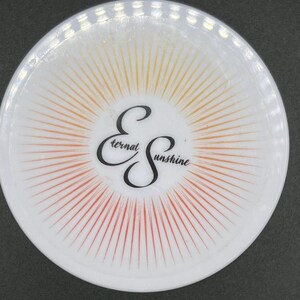 May include: A white, circular coaster featuring a sunburst design in orange and yellow hues. The words "Eternal Sunshine" are written in a stylized script in black.