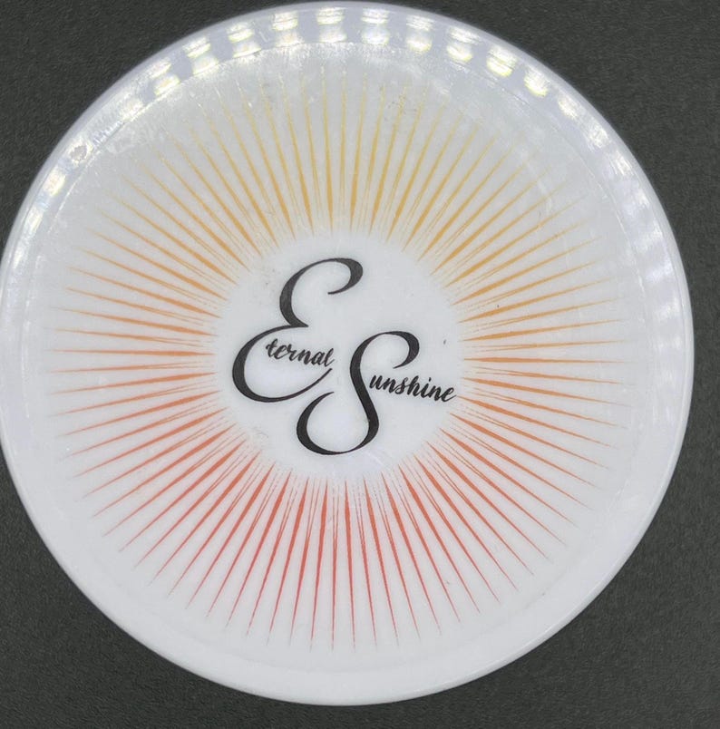 May include: A white, circular coaster featuring a sunburst design in orange and yellow hues. The words "Eternal Sunshine" are written in a stylized script in black.