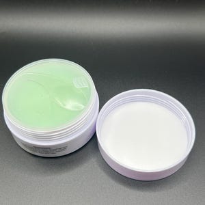 May include: Open white container with a light green gel product inside. The lid is off, revealing the product and the white interior of the container. The product appears to be a skincare item, possibly eye patches.