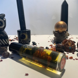 Puede incluir: Un frasco de aceite esencial de vidrio transparente con una tapa negra, lleno de contenido ámbar y verde, se encuentra sobre una superficie blanca. Una figura de búho negro, un portavelas negro y otros objetos decorativos rodean el frasco.