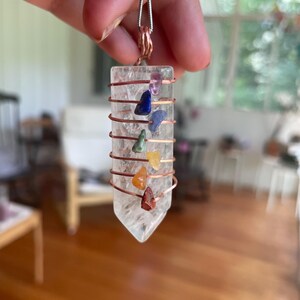 Könnte beinhalten: Ein klarer Quarz-Kristall-Anhänger mit Kupferdrahtwicklung und einer Silberkette. Der Kristall ist mit kleinen, farbigen Edelsteinen in den Farbtönen Lila, Blau, Grün, Gelb, Orange und Rot verziert. Der Anhänger wird von einer Hand gehalten.