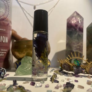 Op de afbeelding: Een heldere glazen roll-on fles met een zwarte dop, gevuld met een vloeistof en paarse bloemen. De fles is omgeven door kristallen en andere decoratieve items. Het algemene thema is spiritueel en holistisch.