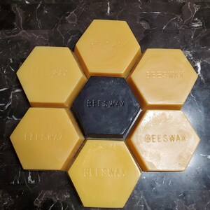 Könnte beinhalten: Ein Wabenmuster aus sechs gelben Bienenwachsblöcken und einem schwarzen Bienenwachsblock. Jeder Block trägt die Aufschrift "BEESWAX".