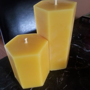 May include: Two yellow hexagonal beeswax candles, one taller than the other, on a black marble surface.