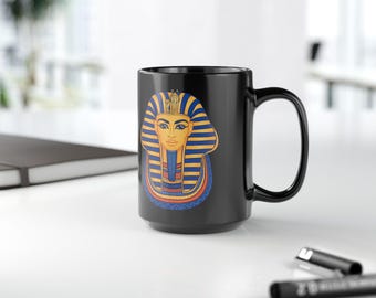 May include: Black ceramic mug featuring a colorful illustration of an Egyptian pharaoh's mask. The design includes blue, yellow, and red accents. The mug has a large handle and is suitable for hot or cold beverages.