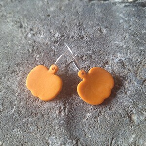 May include: A pair of orange apple-shaped earrings with silver hoops. The earrings are made of a solid, opaque material and have a smooth surface. The earrings are displayed on a textured grey surface.