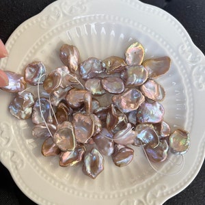 May include: A collection of iridescent, pearlescent, and pink freshwater pearls in various shapes and sizes. The pearls are arranged on a white plate with a decorative border.