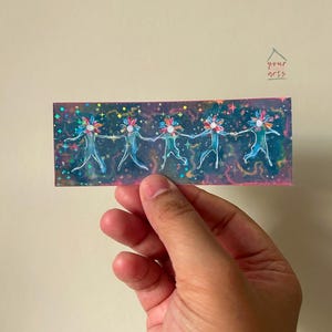 Peut inclure: Un autocollant holographique représentant cinq personnages stylisés se tenant par la main. Chaque personnage a une tête en forme de fleur et est représenté dans des tons de bleu et de turquoise. Le fond est pailleté et le texte "your arts" est inclus.