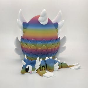 May include: A 3D printed rainbow-colored egg with white wings and a textured scale pattern, accompanied by a small, articulated dragon figure featuring a matching rainbow gradient body, white wings, and large black eyes with pink cheek accents.