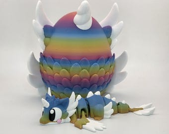May include: A 3D printed rainbow-colored egg with white wings and a textured scale pattern, accompanied by a small, articulated dragon figure featuring a matching rainbow gradient body, white wings, and large black eyes with pink cheek accents.
