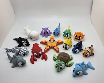 May include: A collection of small, colorful, stylized sea creature figurines including a white whale, purple octopus, orange starfish, red lobster, green sea turtle, blue dolphin, and various tropical fish with large eyes and smooth surfaces.