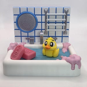 May include: A miniature bathroom scene featuring a yellow duck in a bathtub with pink soap and bubbles. The background has a blue and white tiled wall with a mirror and a towel rack. The soap bars are pink and have the word "SOAP" printed on them.