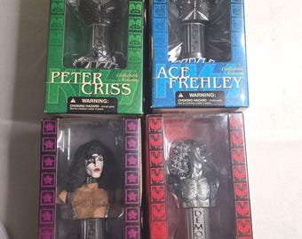 May include: Four collectible statuettes of KISS band members in their signature makeup and costumes. Each bust is displayed in a box with the band member's name: Peter Criss, Ace Frehley, Starchild, and Demon. The boxes have warning labels.