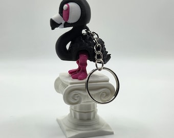 May include: A black flamingo keychain with pink feet and eye accents, perched atop a white decorative column. The keychain has a silver ring for attaching keys. The flamingo has a unique, stylized design.