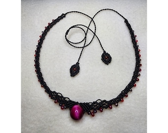May include: A black macrame necklace with red and purple beads. The necklace features a large, round, purple gemstone pendant. The necklace has two long, dangling cords with decorative ends. The necklace is displayed on a white surface.