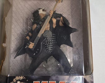 May include: A figurine of a rock musician in a black studded outfit, playing a black bass guitar. The figure has white face paint and long black hair. The figurine is in a clear plastic display case.