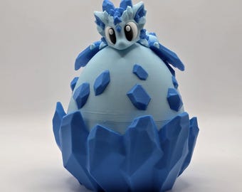 May include: A light blue and dark blue 3D-printed dragon figurine emerging from a light blue egg. The egg is decorated with dark blue geometric shapes. The dragon has large eyes and spiky features.