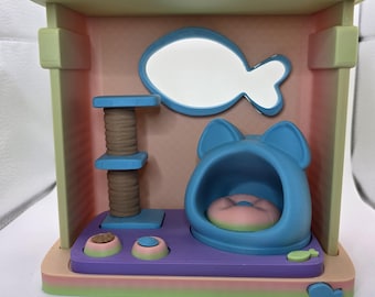 May include: A colorful cat house toy with a blue cat bed, a scratching post, and a fish-shaped window. The house is decorated with pastel colors, including pink, green, and purple. A small fish-shaped decoration is on the top.