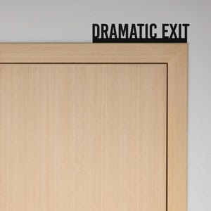 May include: A wooden door with a black sign above it that reads "Dramatic Exit".