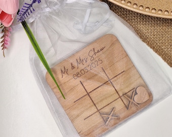 May include: A wooden tic-tac-toe game with the text "Mr & Mrs Shaw 08.03.2025" engraved on the surface. The game is in a white organza bag.