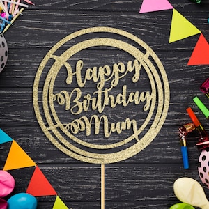 Peut inclure: Un cake topper doré à paillettes "Happy Birthday Mum" dans un design circulaire, entouré de décorations de fête. Les décorations comprennent des ballons, des fanions colorés, des serpentins et des bougies, le tout sur une surface en bois sombre.