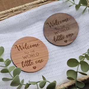 May include: Two round wooden plaques with the text "Welcome to the world little one" engraved on them. The plaques are on a white blanket with green leaves in the background.
