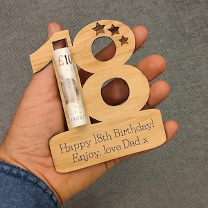 May include: Wooden "18" birthday gift with a rolled-up £10 bill. The number "18" is cut out of wood, with three star cutouts above the "8". The base reads "Happy 18th Birthday! Enjoy, love Dad x".