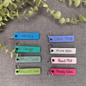 Könnte beinhalten: Acht rechteckige Holzschlüsselanhänger in verschiedenen Farben wie Blau, Grün, Türkis, Hellgrün, Lila, Weiß, Pink und Magenta. Jeder Anhänger hat einen Metallring und eine schwarze Gravur mit den Texten Office, Garage, Man Cave, Grandad's Shed, Cake Shed, Front Door, Beach Hut und Beauty Salon.
