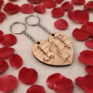 May include: Two wooden heart-shaped keychains with silver chains and keyrings. The keychains are engraved with the names "Simon" and "Denise" and the date "12.03.2024". Red rose petals surround the keychains.