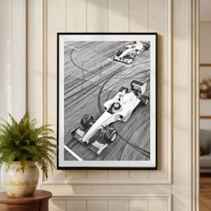 May include: A black and white framed wall art print featuring an aerial view of two Formula One race cars on a track. The cars are positioned on asphalt marked with dark tire skid marks, showcasing the aerodynamic design of the racing vehicles.