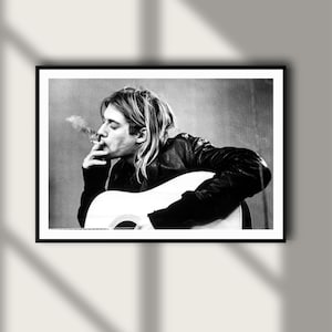May include: A black and white photographic print featuring a person with shoulder-length hair wearing a dark jacket, holding a cigarette to their lips with smoke rising, while playing an acoustic guitar. The image is framed in a thin black border.