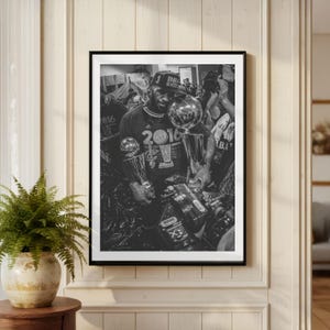 May include: A black and white framed photograph depicting a basketball player wearing a 2016 NBA Champions cap and t-shirt, holding a large silver trophy. The player is surrounded by cameras and equipment in a crowded, celebratory locker room setting.