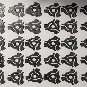 May include: A collection of forty-five black plastic 45 RPM record adapters arranged in a grid on a white sheet. Each adapter features a circular center hole and three curved arms extending outward, designed to fit standard vinyl records.