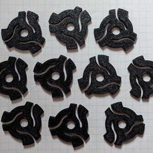Puede incluir: Una colección de diez adaptadores negros para discos de 45 RPM, cada uno con una superficie texturizada y un diseño distintivo de tres puntas. Los adaptadores están dispuestos sobre un fondo de cuadrícula blanca, mostrando su forma y detalle.