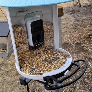 May include: A white bird feeder with a clear plastic cover over a black and white camera. The feeder is filled with birdseed and has a black handle on the side. The feeder is labeled "Bird Buddy".