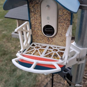 May include: A white bird feeder with a blue roof and a white camera inside. The feeder is mounted on a metal pole and has a red and white platform.