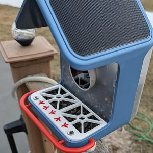 May include: A blue and white bird feeder with a red perch. The feeder has a black textured roof and a clear plastic container to hold birdseed. The perch has red bird footprints and a red metal support.